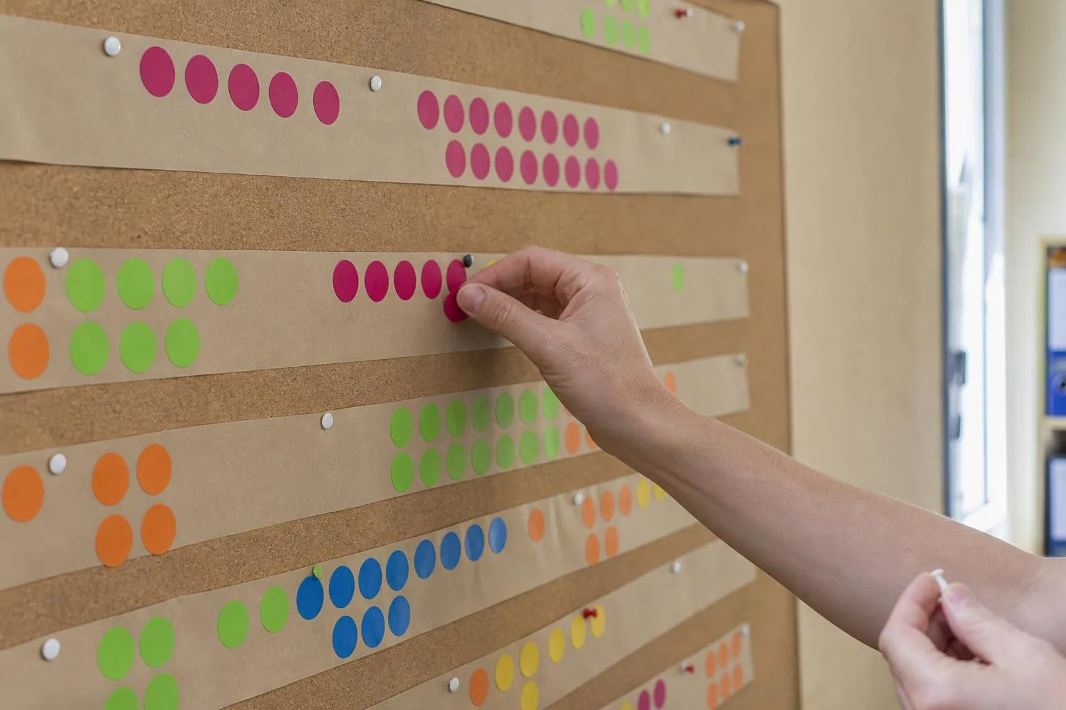 Un mur de bureau couvert d’un planning annuel fait de bandes de papier kraft et de pastilles de couleur, une main déplaçant une pastille