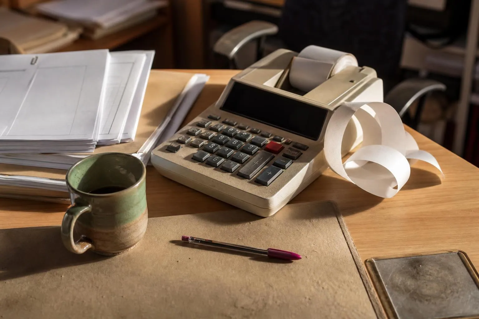 Une calculatrice à bandelette posée sur un bureau, son ruban de papier déroulé en boucle à côté d’une pile de feuilles et d’une tasse